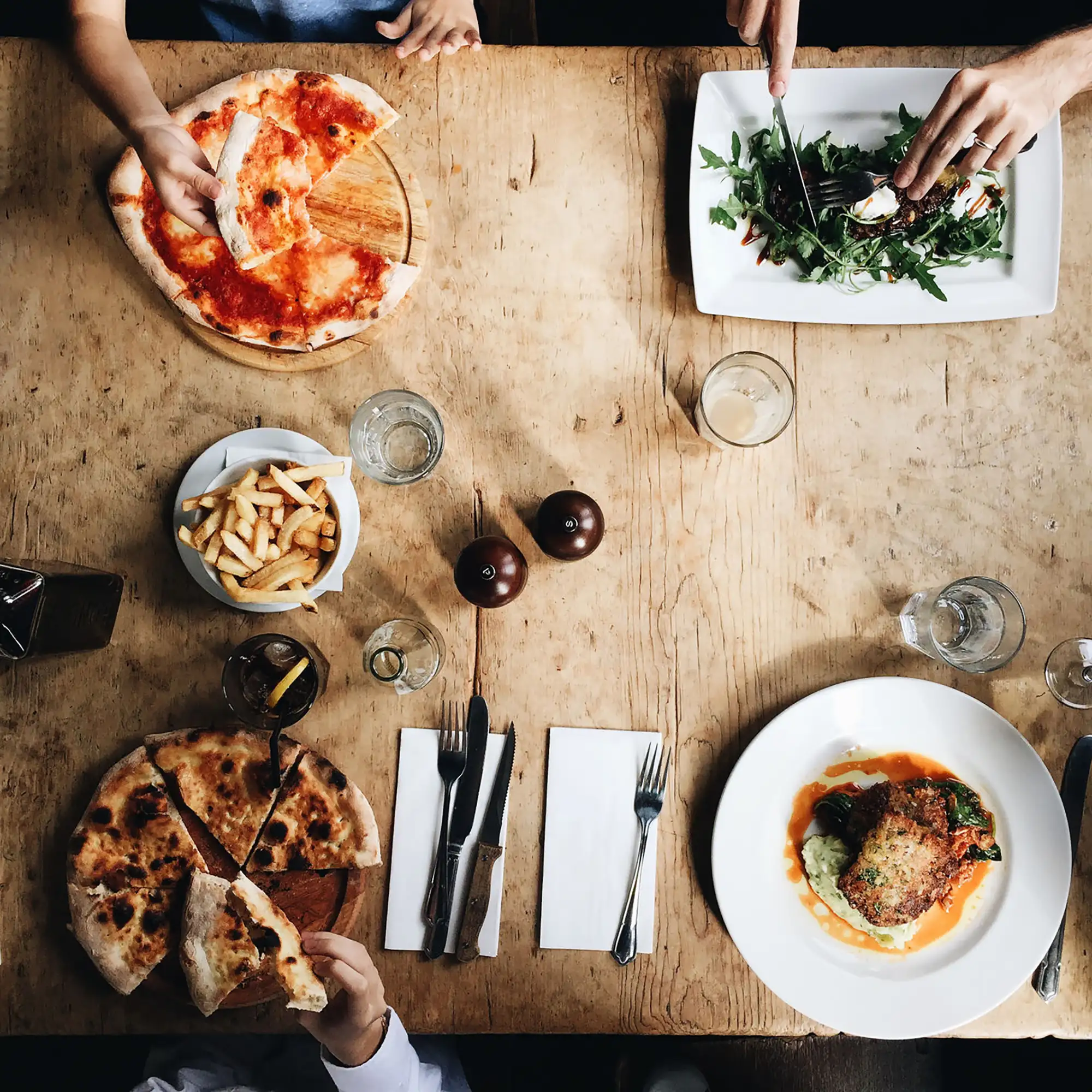 Overhead view of a rustic wooden table with hands reaching for pizza, fries, and other dishes during a meal.
