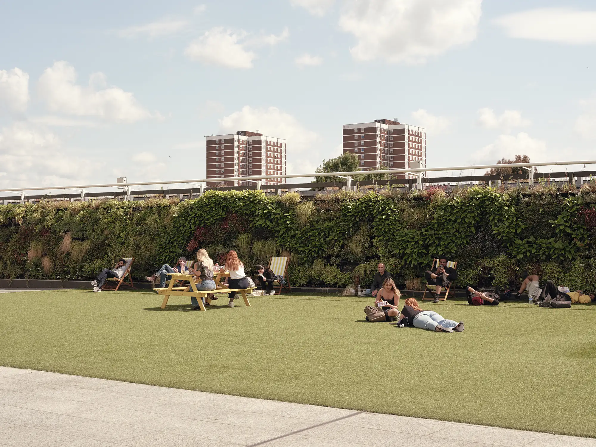 People relaxing on artificial green grass with picnic benches and residential buildings in the background.