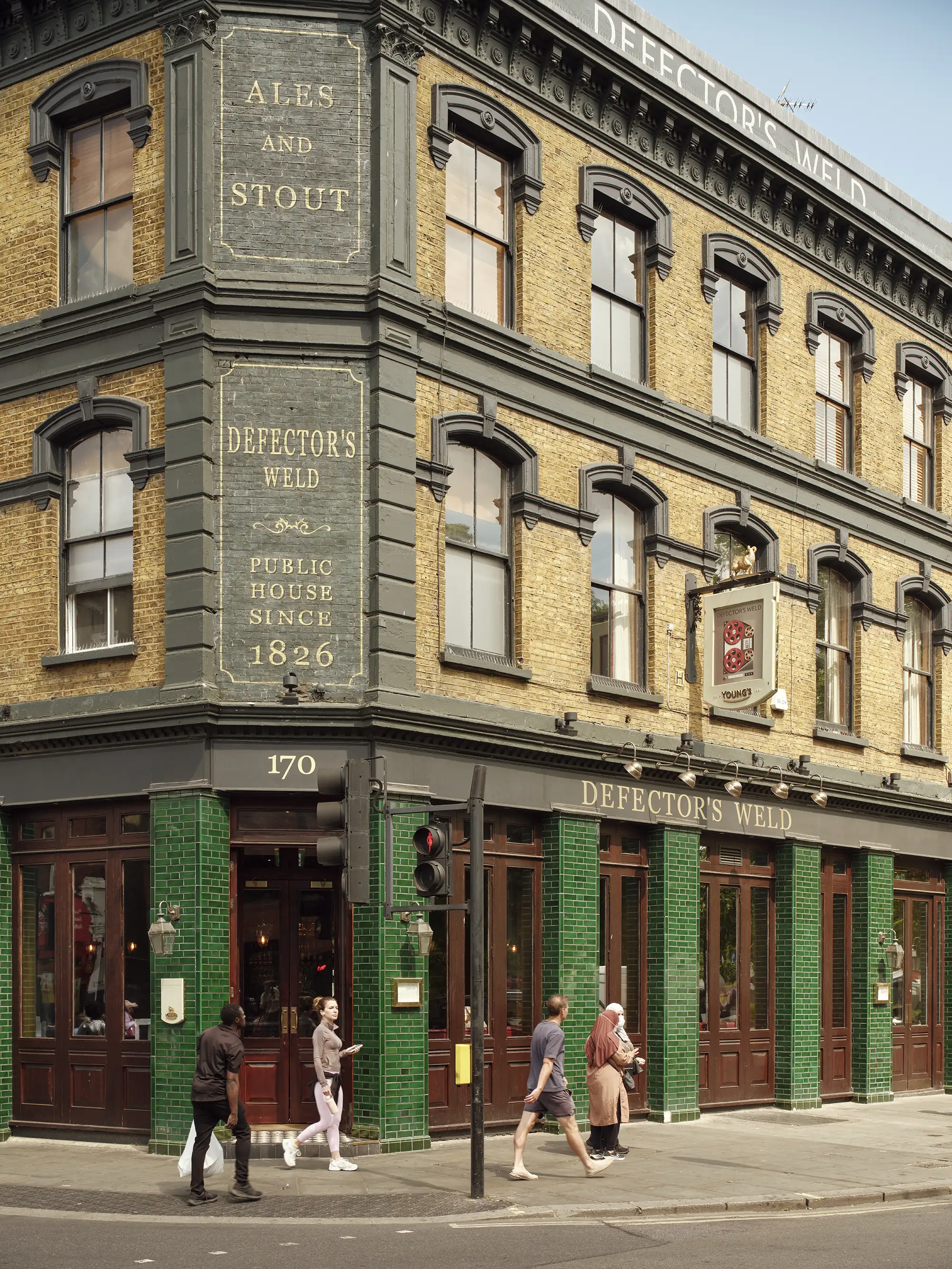 The exterior of Defector's Weld pub with classic brickwork, dark green tiling, and pedestrians walking by.