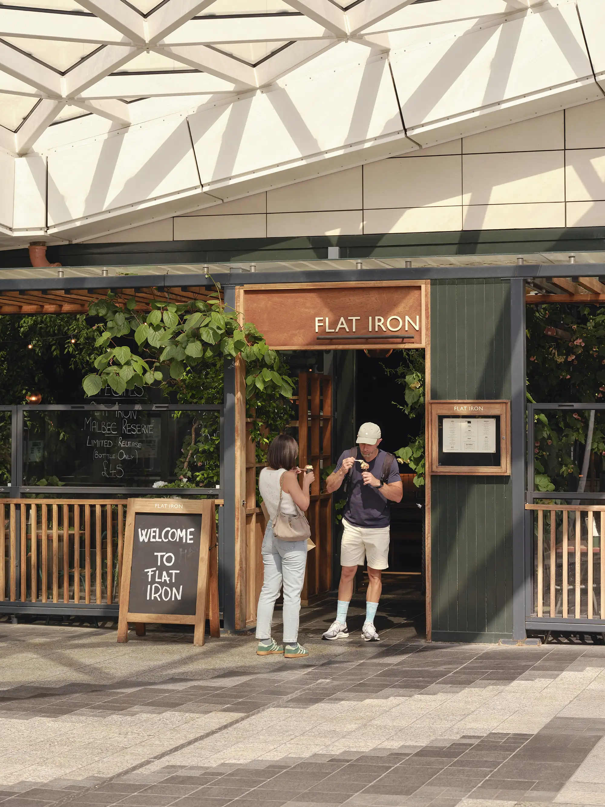 A man and woman standing outside the Flat Iron restaurant entrance, sharing food.