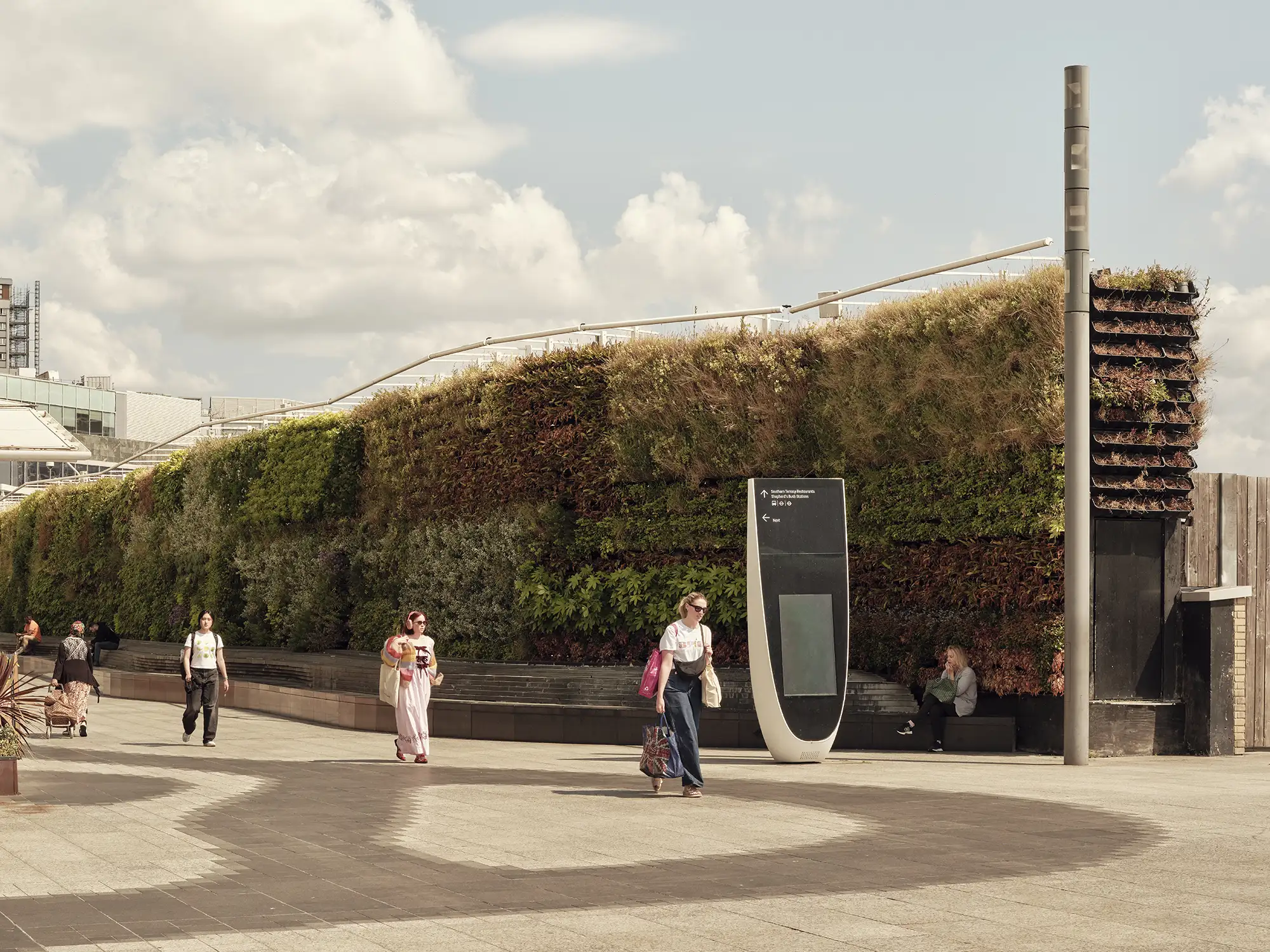People walk past a large, lush green living wall under a cloudy sky.