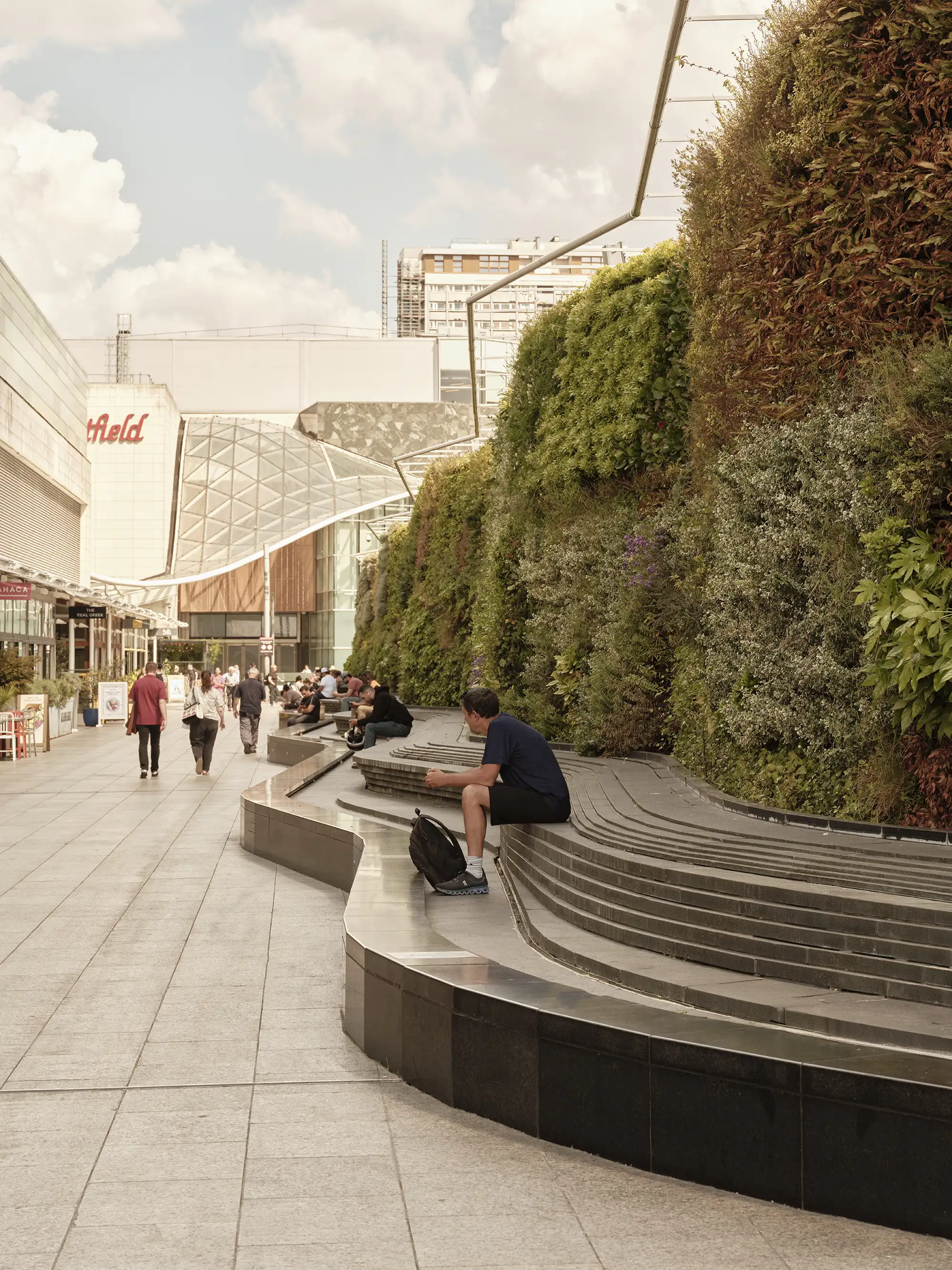 A man sits on curved steps beside a vibrant green living wall, with shoppers and buildings in the background.