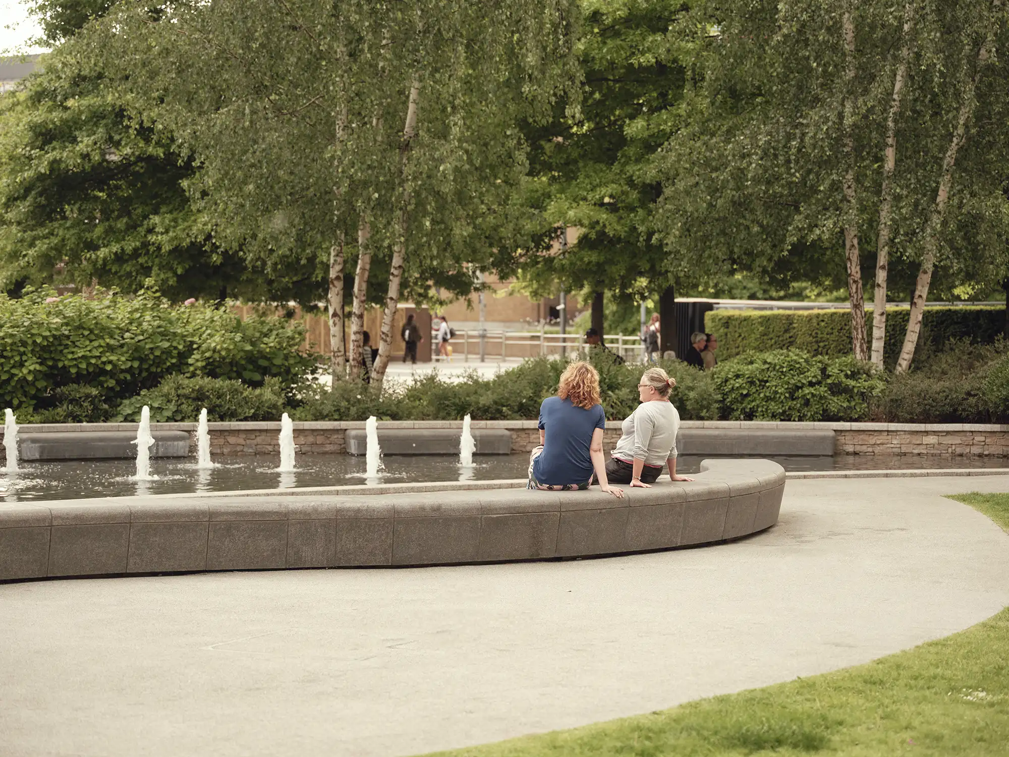 Relaxing scene in an urban park with a curved stone bench, water fountains, and lush green foliage.